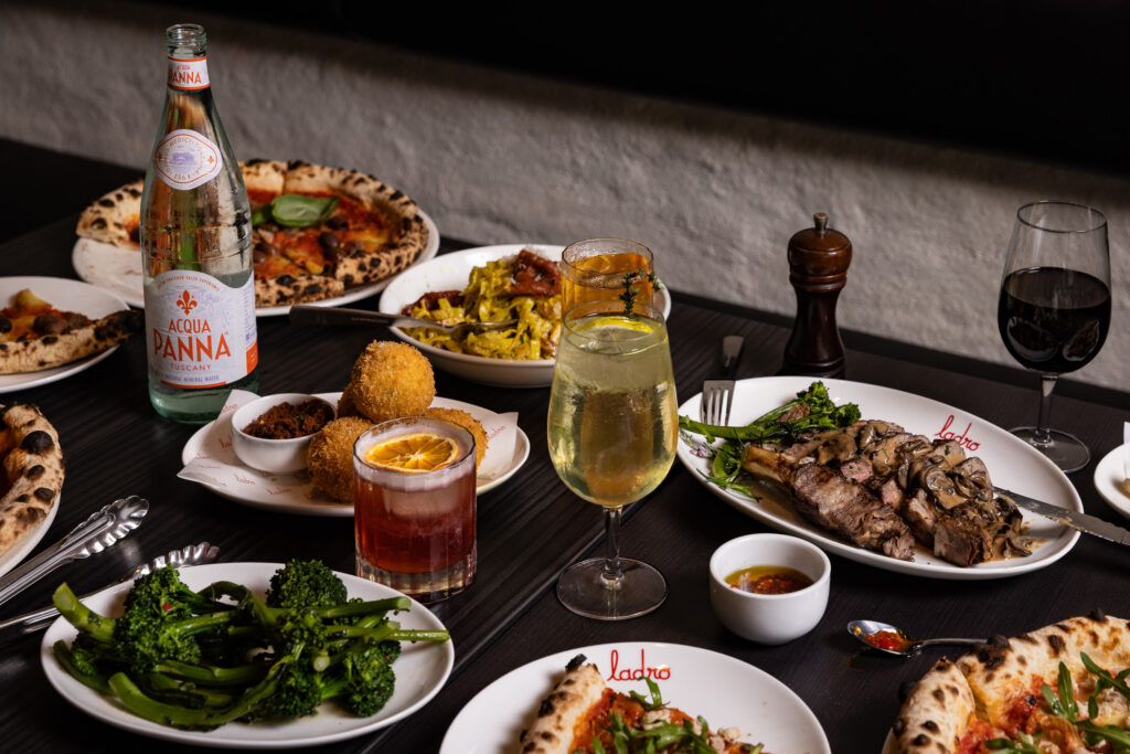 A bunch of Italian dishes on a table; including arancini, pizza and limoncello spritz.
